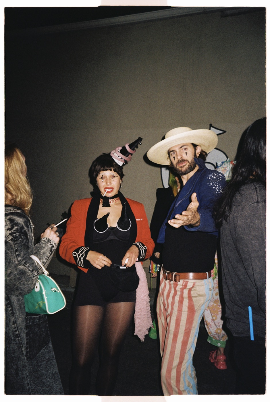 Neeff in a wide-brimmed hat, painted tear marks, and red-and-white striped trousers, gesturing toward the camera at a costumed night gathering