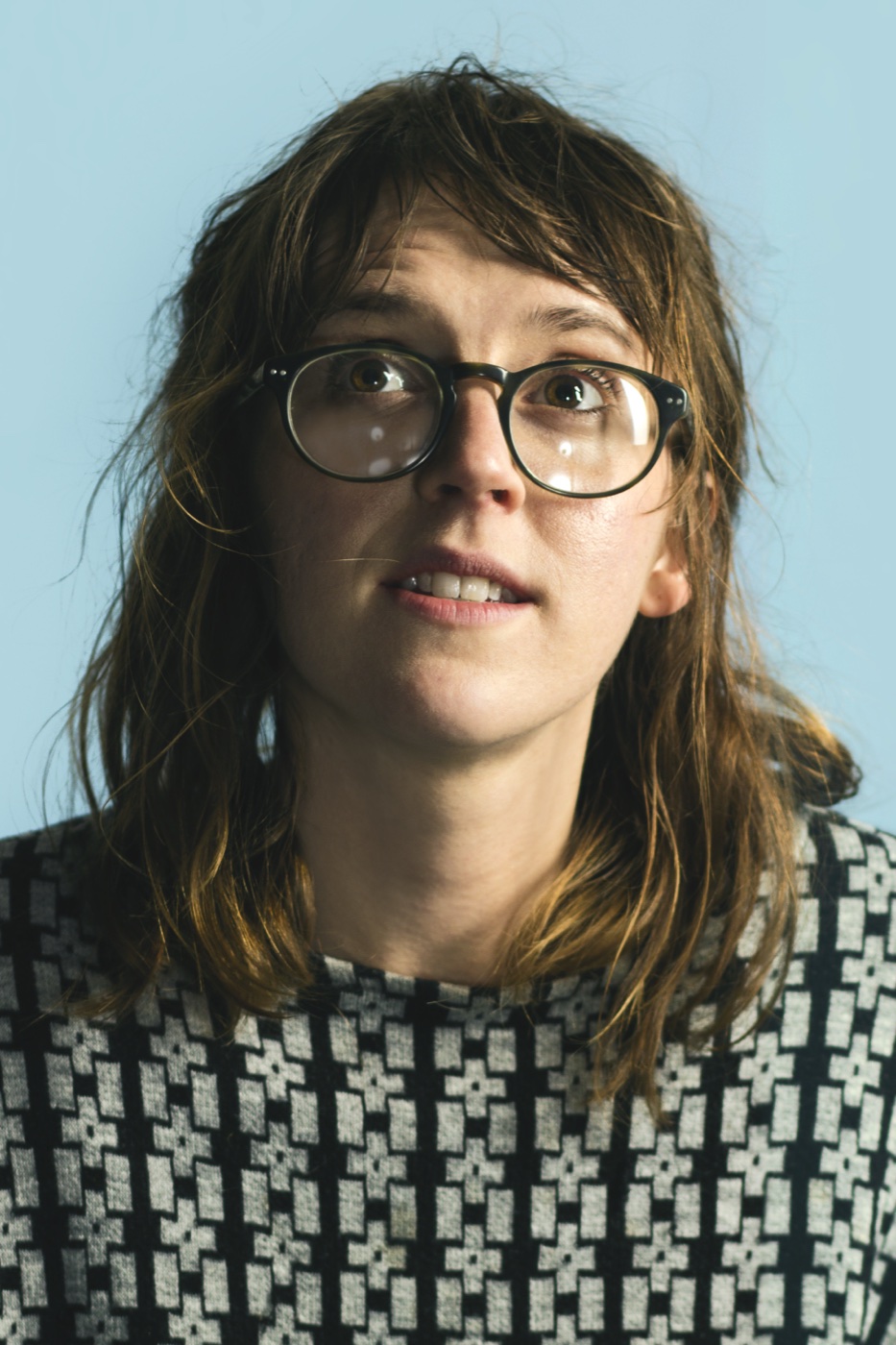 Photographic portrait of a person with shoulder-length hair and round glasses, looking up against a pale blue wall, wearing a black-and-white patterned sweater