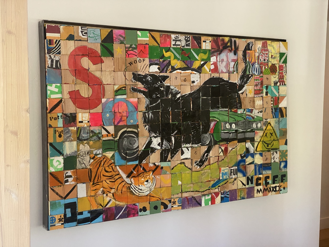 A framed painting made of hundreds of small painted wooden cubes, showing a black dog leaping over a green car, a tiger, a red letter S, and a domed cathedral, signed NEEFF MMXXI